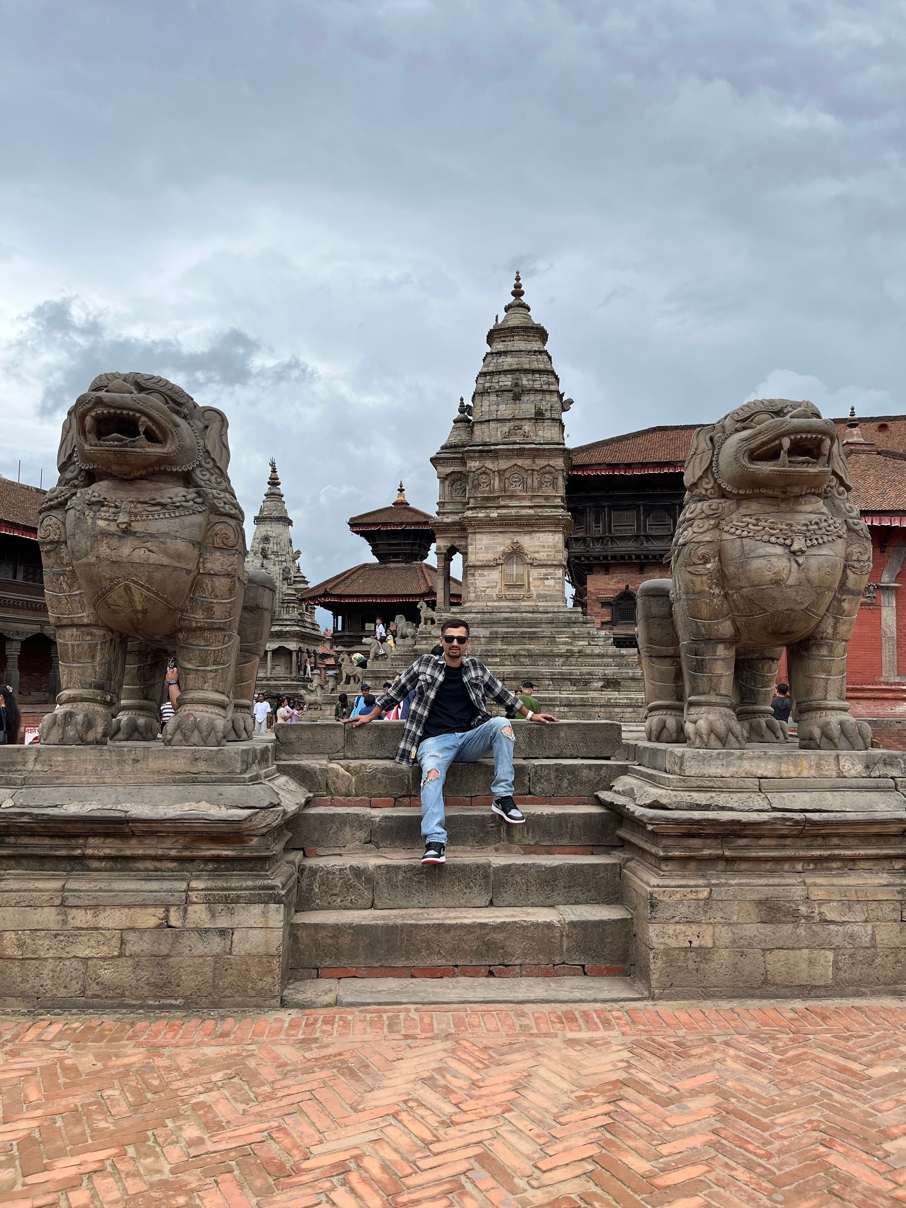 Person posing at Bhaktapur Durbar Square with stone lion statues and traditional architecture.