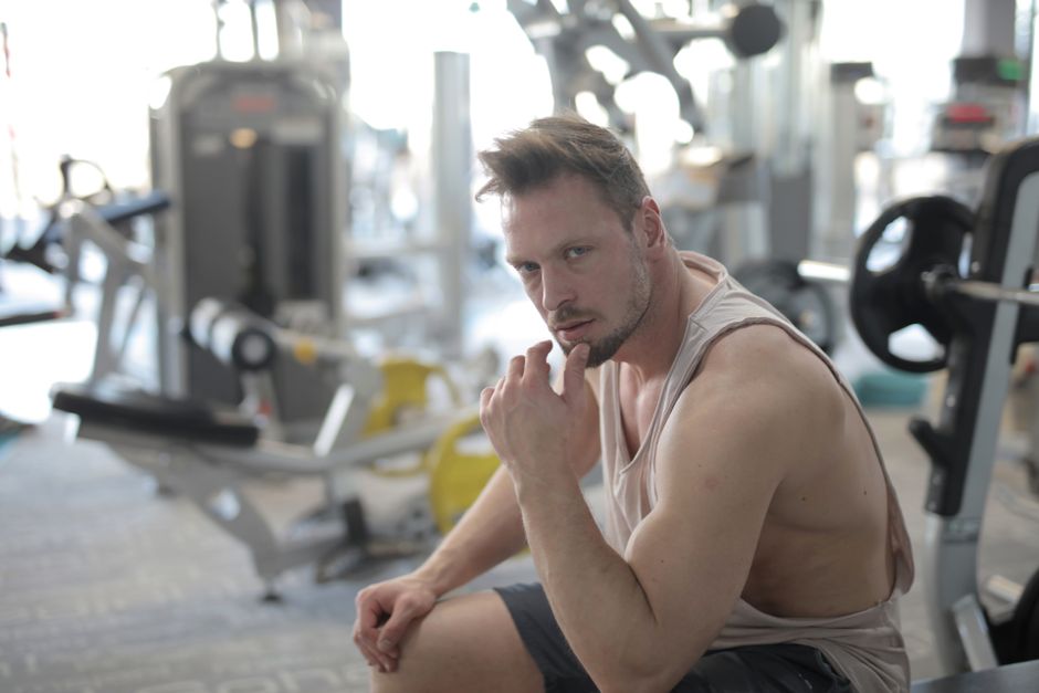 Confident young sportsman in sportswear sitting on bench and resting after workout in modern fitness center and looking at camera