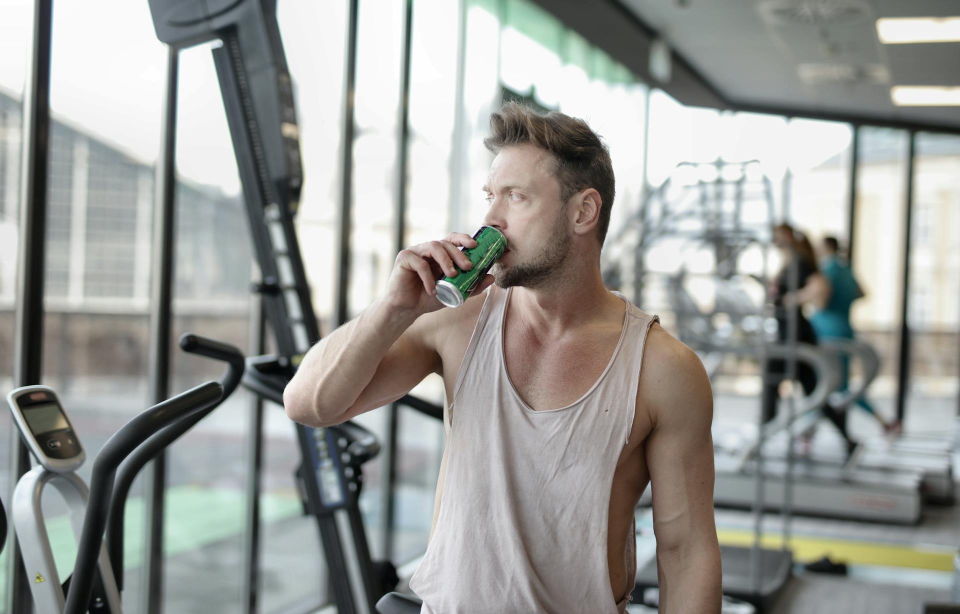 Strong muscular young male athlete in casual sportswear drinking fresh cold energy drink while resting after intense workout and looking away through window in gym