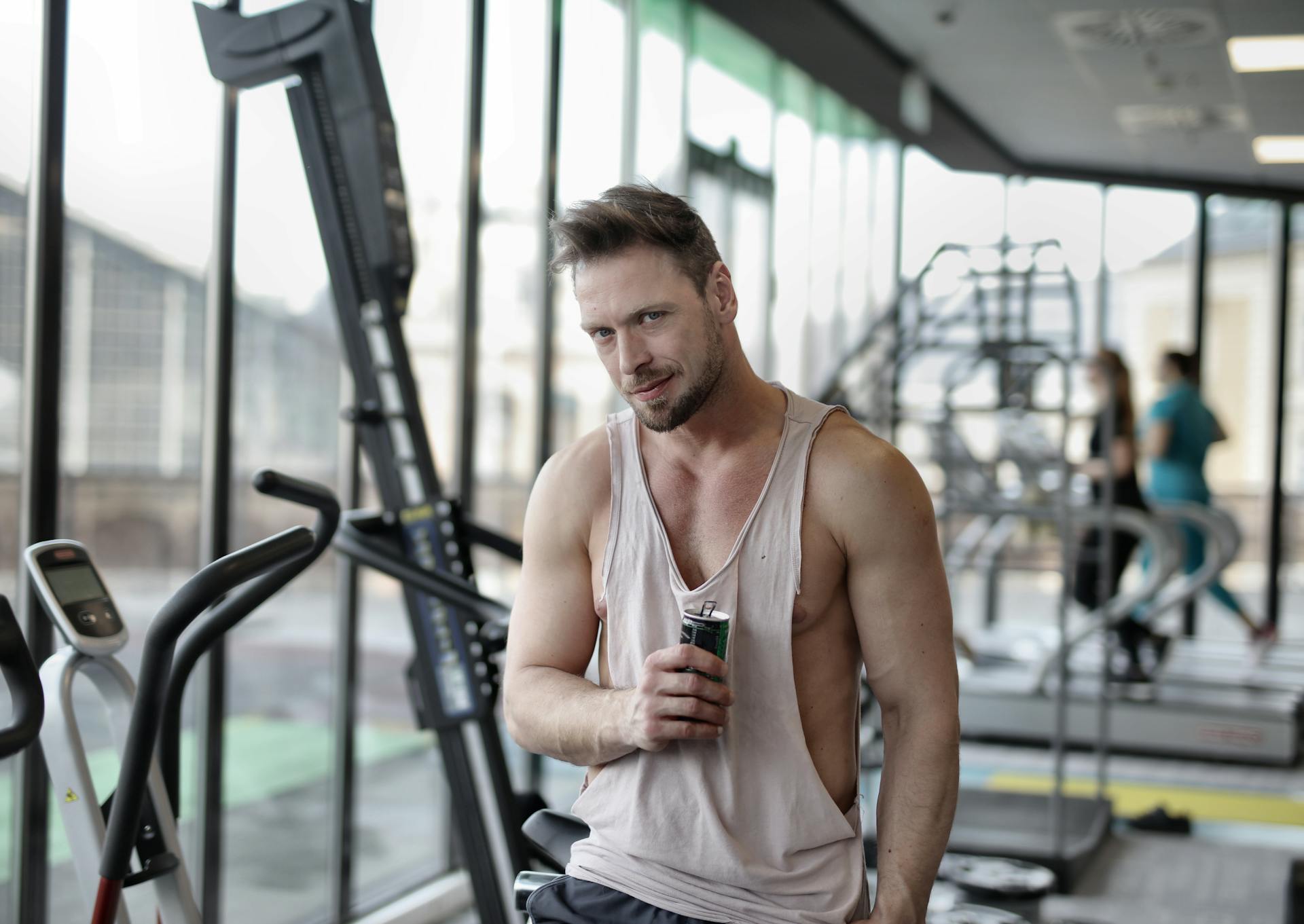 Positive young muscular bearded male athlete in casual sportswear standing near window in modern gym with bottle of cold energy drink and looking at camera