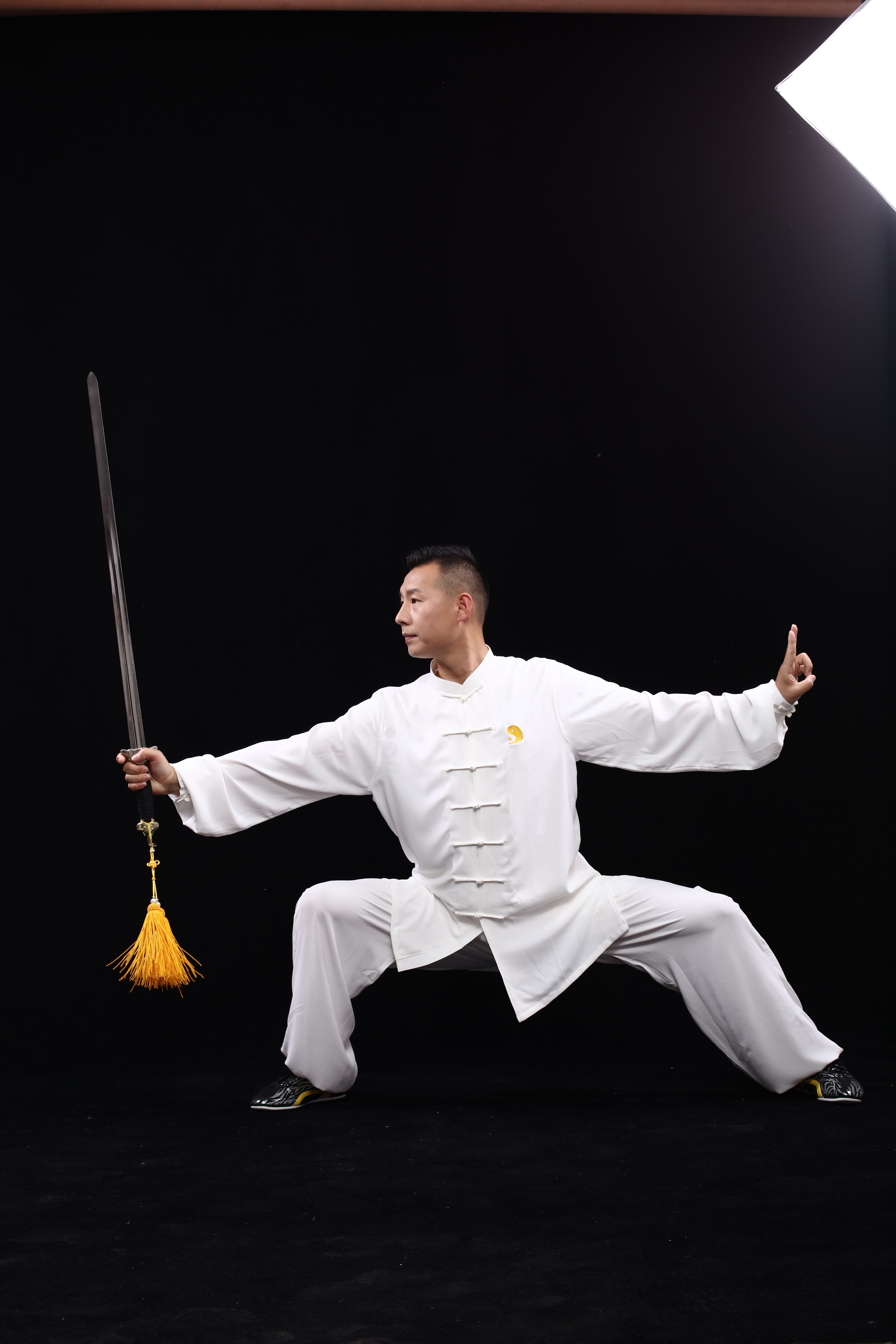 Martial artist demonstrates traditional pose with sword in full white uniform against a dark backdrop.
