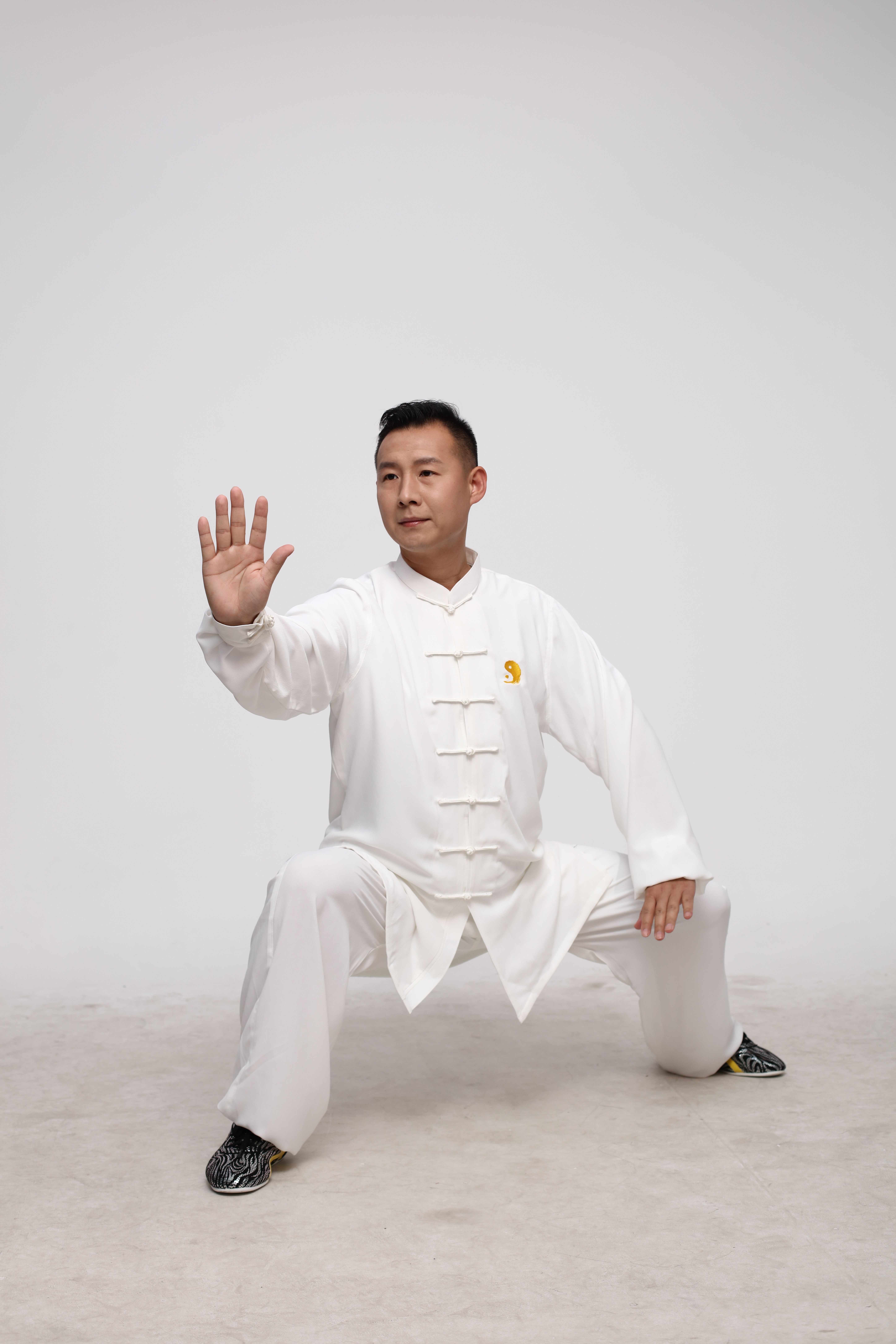 An adult male in a Tai Chi pose, wearing a classic white outfit, indoors.