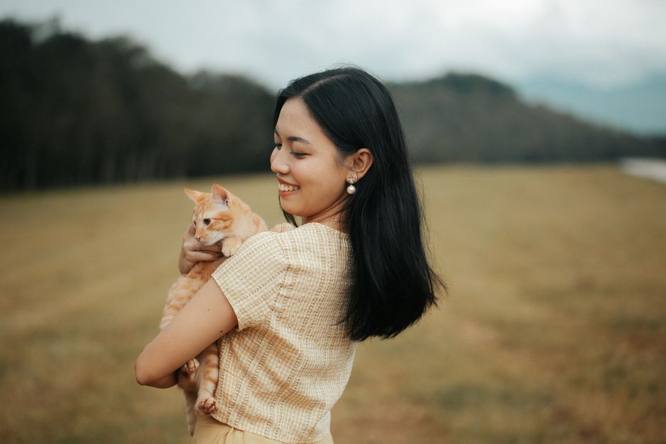 Smiling woman holding a ginger cat in a lush field, conveying warmth and companionship.