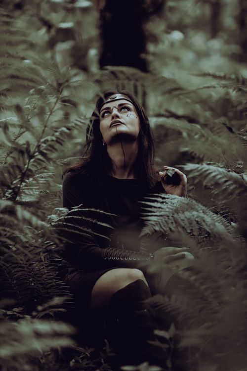 Free A woman in a mystical forest setting gazes upwards, surrounded by ferns and dark foliage. Stock Photo