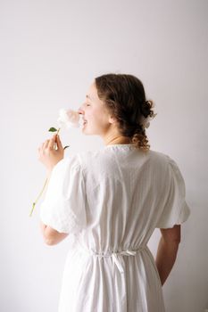 A woman in a white dress holding a white rose, enjoying its scent indoors.