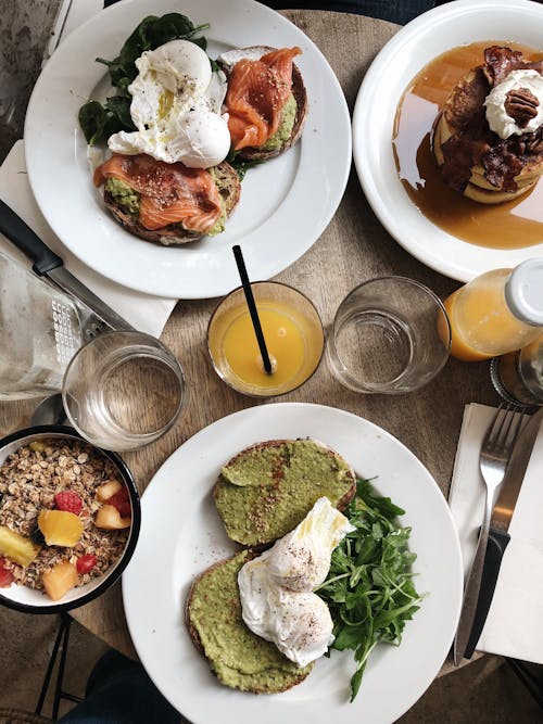 Top view of plates of assorted healthy toasts with poached eggs and natural juice served on wooden table near oatmeal bowl and sweet pancakes