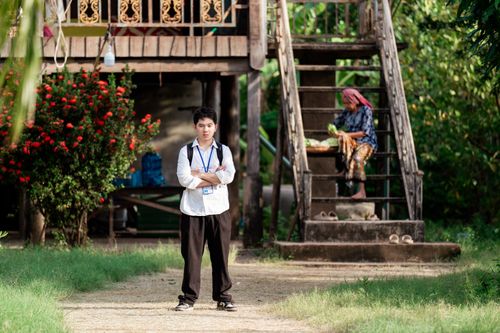 Free A young student stands confidently in front of a rural home with traditional wooden architecture. Stock Photo