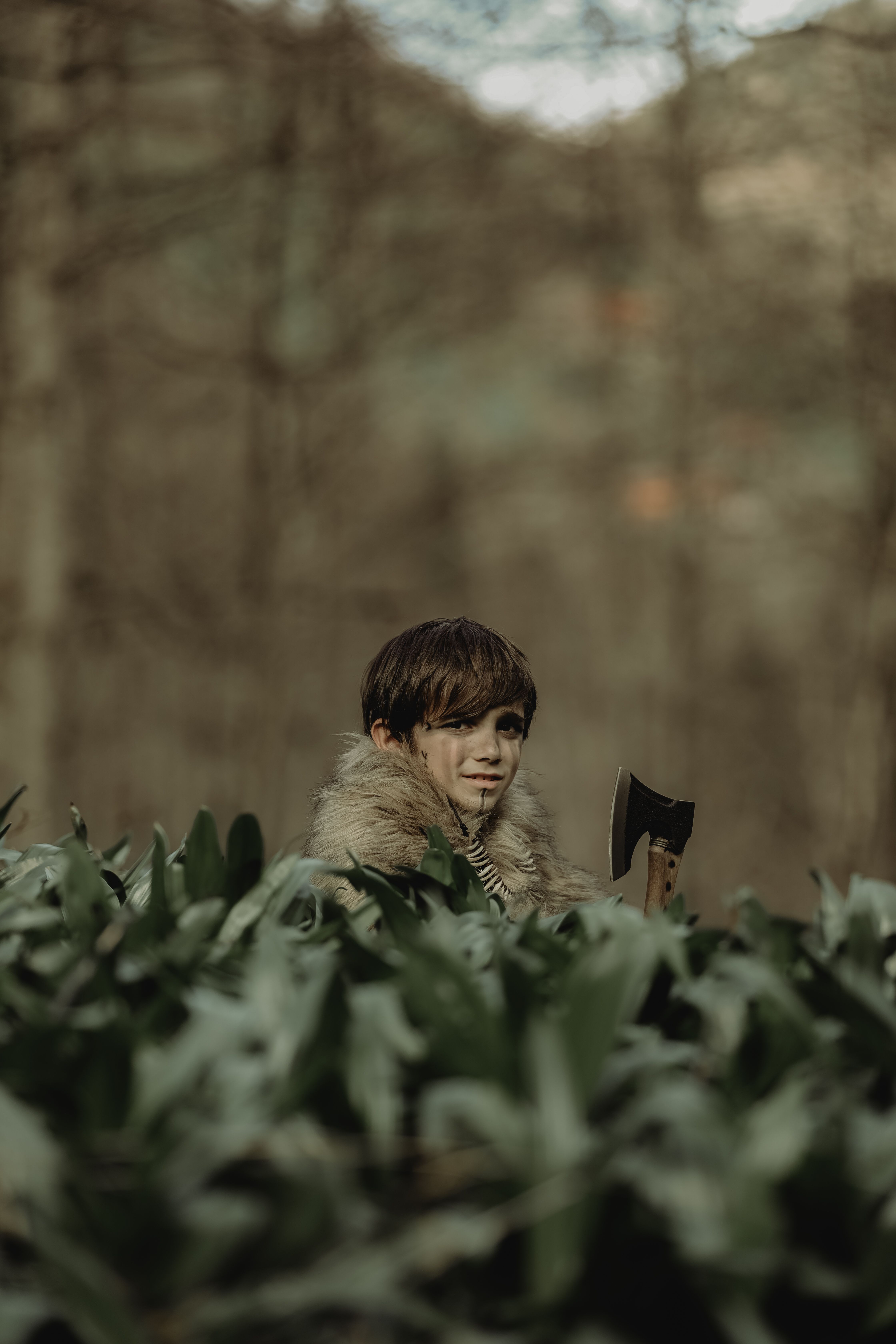 A child dressed in a fur coat holds an axe amidst a lush forest setting.