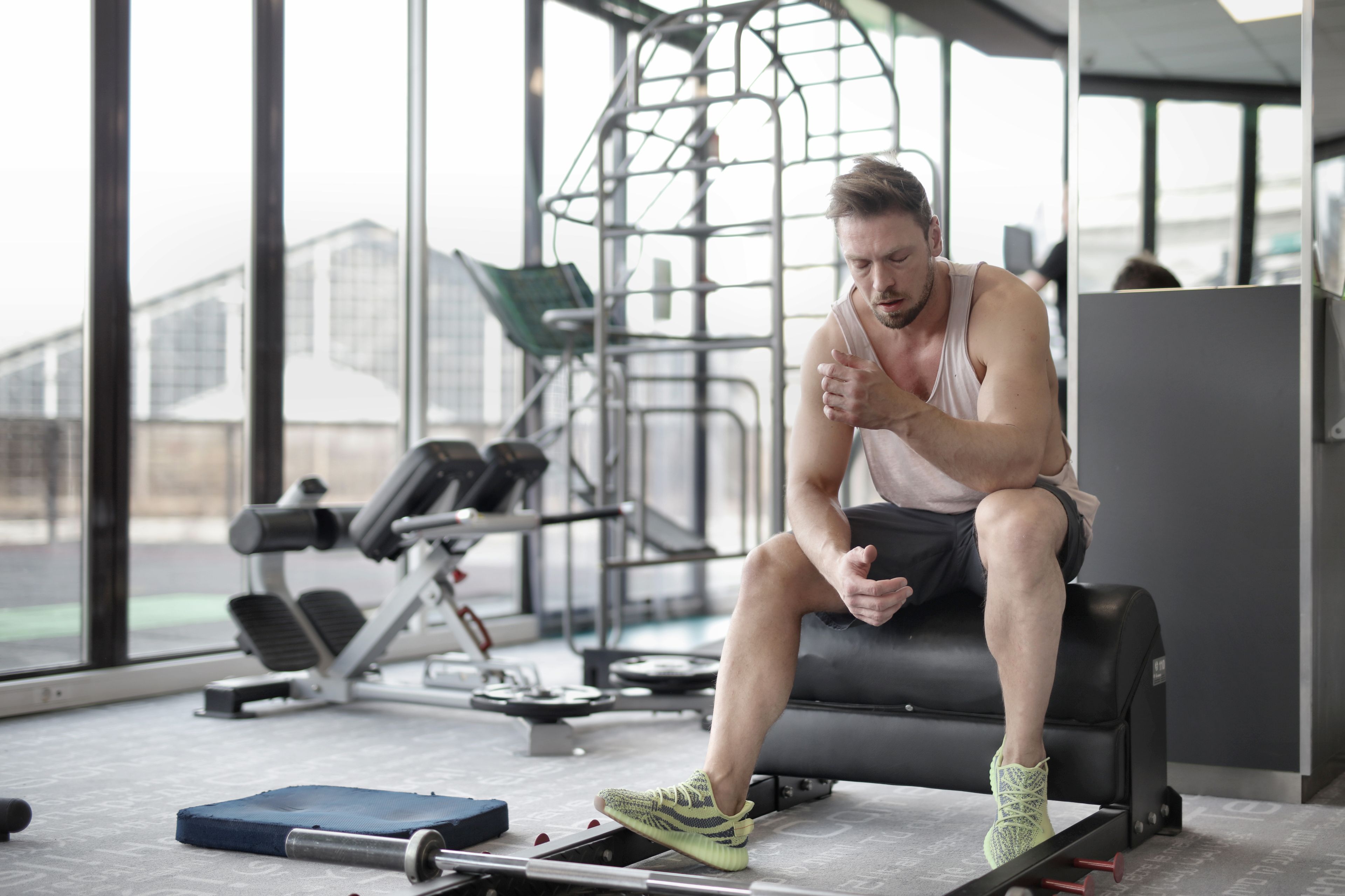Tired young muscular male athlete in casual sportswear and sneakers sitting on bench and resting after intense weightlifting workout in modern fitness club