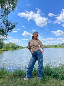 A young woman stands confidently by a serene lake under a bright summer sky.