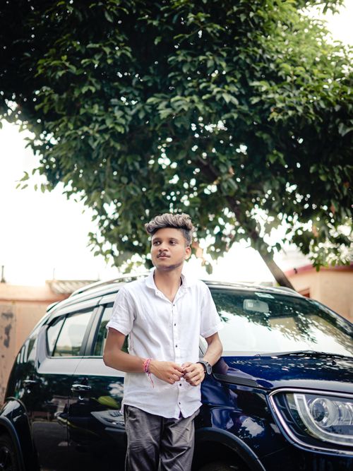 Free A young man in a white shirt leaning against a car under a tree, enjoying the outdoors. Stock Photo
