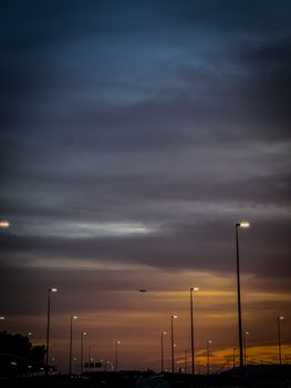 A plane flies over street lamps during a vibrant sunset in Spain, highlighting contrasting skies.