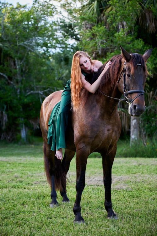 Free A woman in a green dress relaxes on a brown horse in a lush outdoor setting. Stock Photo