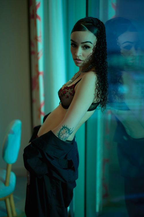 Free Elegant woman posing indoors with moody lighting, wearing fashionable attire and standing by a window. Stock Photo