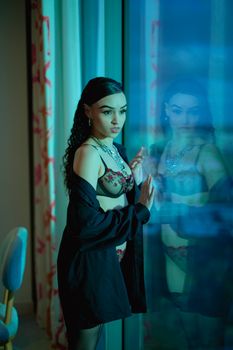 A stylish woman in lingerie and a shirt gazes thoughtfully out a window with soft lighting.