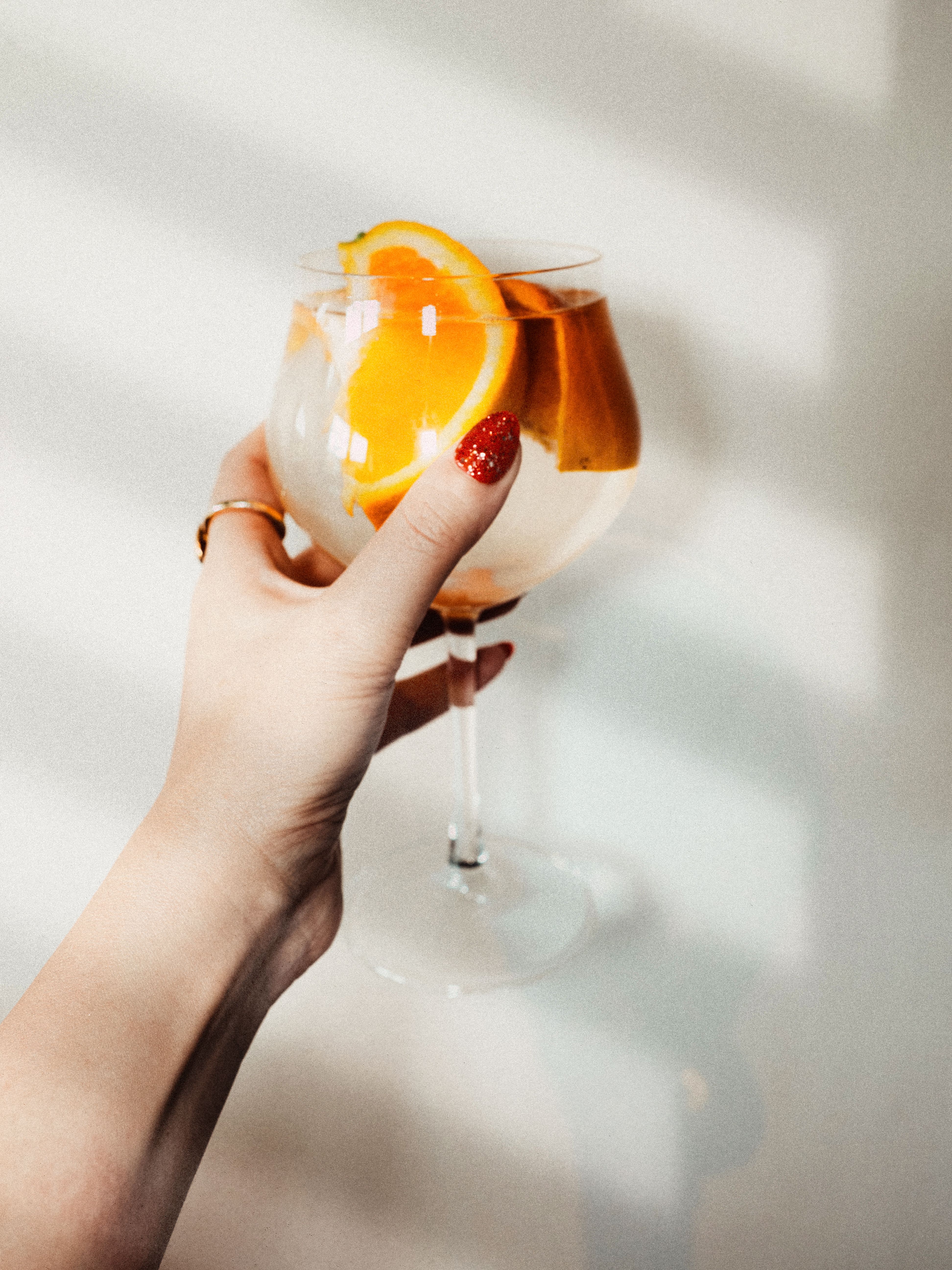 A woman's hand with red nails holding a cocktail glass with orange slices. Light shadow background.