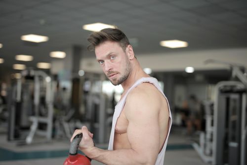 Free Side view of strong serious bearded male athlete in sportswear standing in modern fitness center doing kettlebell bicep curls and looking at camera Stock Photo