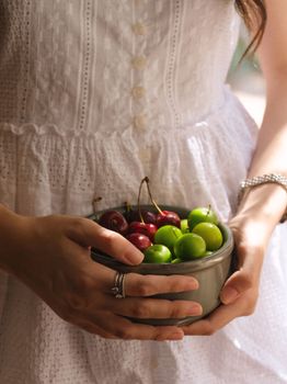 Close-up of a woman in a white dress holding a bowl of fresh cherries and green plums.
