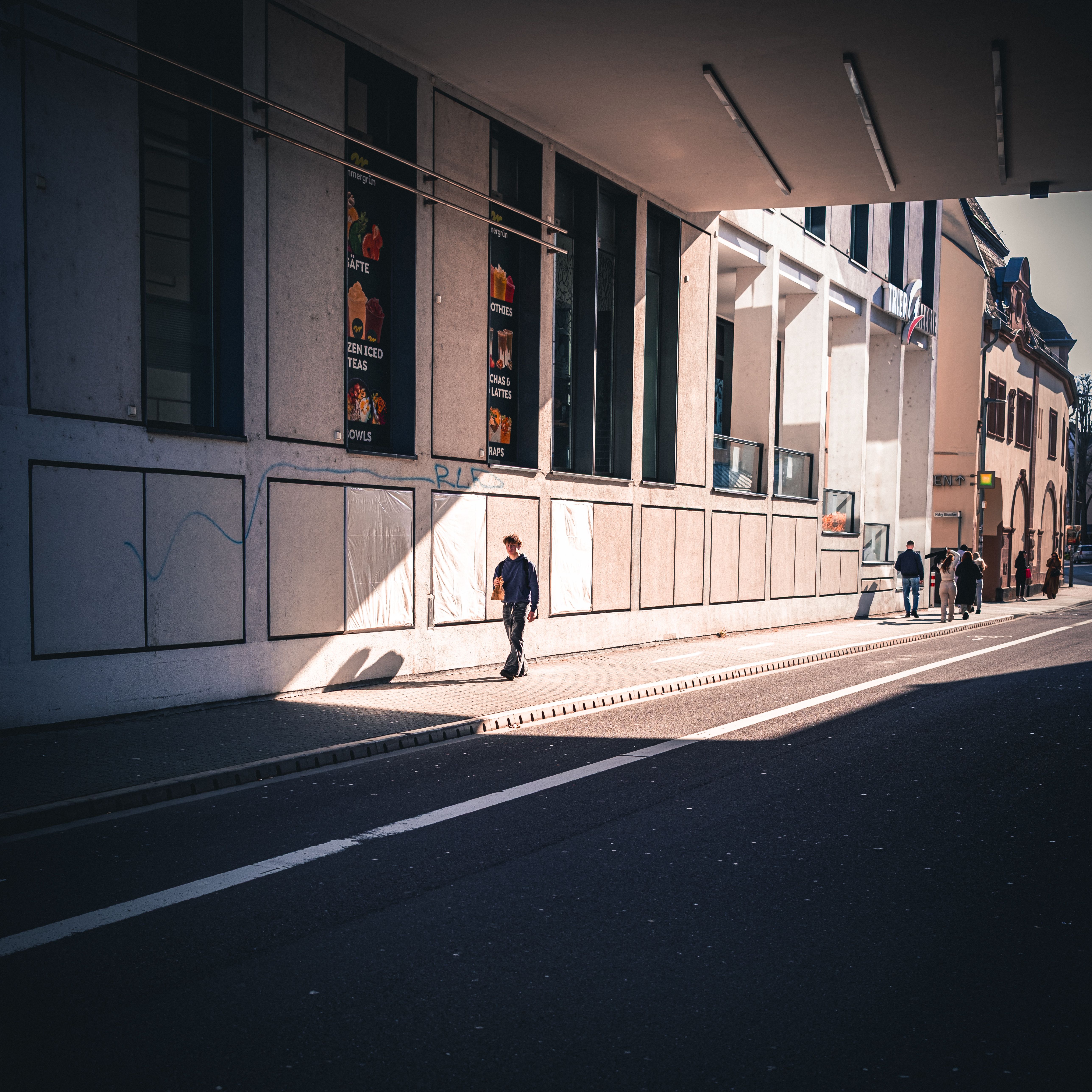 Street view under bridge with people and sunlit shadows on the sidewalk.
