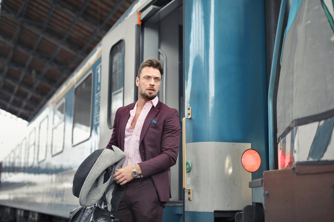 Serious bearded male passenger in stylish suit standing near train with travel bag and coat in hand and looking away