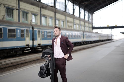 Serious stylish businessman in elegant white shirt and purple jacket holding leather bag and coat in hand standing on platform on railway station and waiting for train