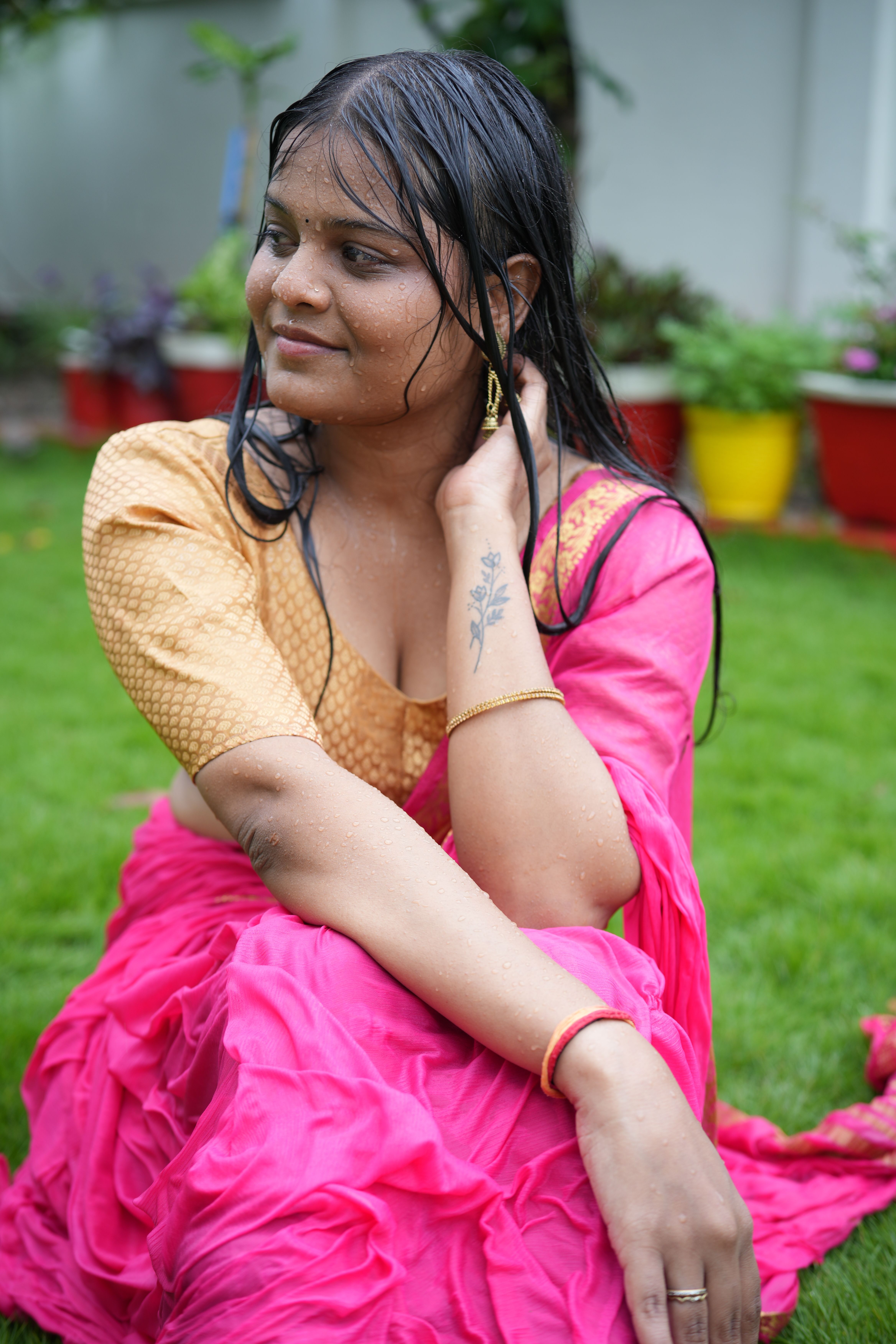 A woman in a vibrant saree sits on a lawn after rain, surrounded by colorful pots.
