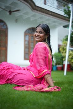 A woman in a vibrant pink saree enjoys a rainy day outdoors, smiling and sitting on green grass.