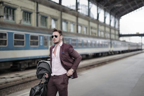 Confident trendy male traveler in sunglasses standing on platform on railway station