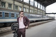 Confident trendy male traveler standing on railroad platform with coat and travel bag