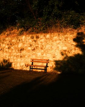 An empty bench casts long shadows against a sunlit stone wall, evoking a serene, contemplative mood.