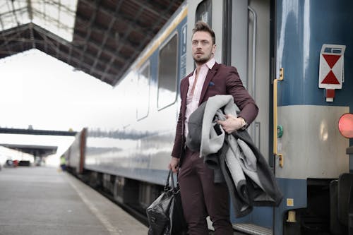 Serious stylish bearded businessman in trendy suit holding bag and coat in hands standing near train on platform in railway station and looking away