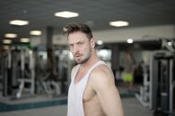 Serious young bearded male athlete in casual sportswear standing in modern fitness center while resting after workout and looking at camera