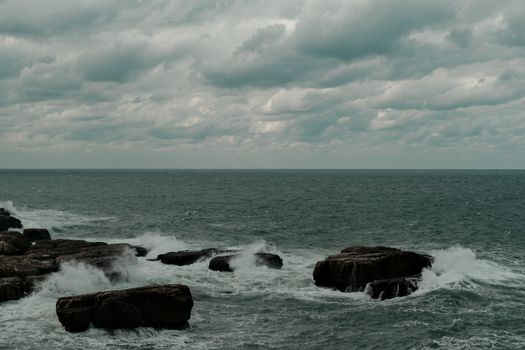 Stormy sea waves crash against rugged rocks in İstanbul, Türkiye, under an overcast sky.