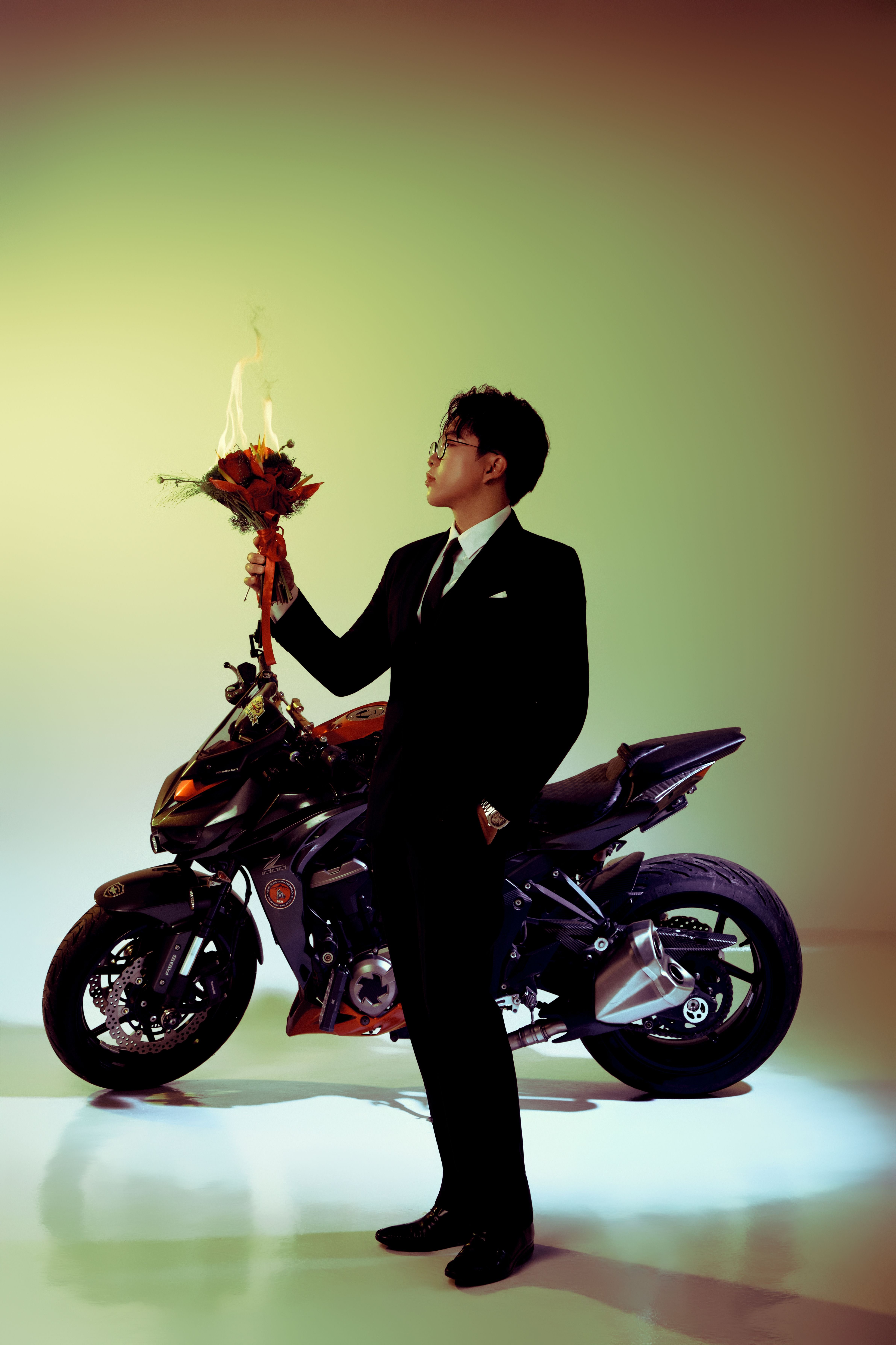 A man in a suit holds a bouquet of flaming roses near a motorcycle under dramatic lighting.