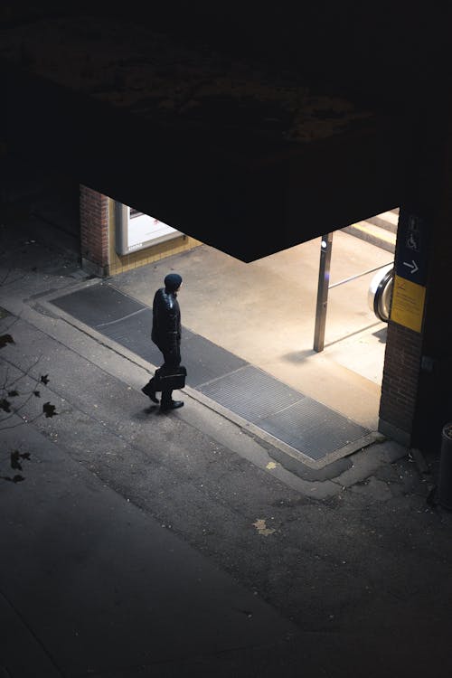From above of anonymous male in warm clothes and hat walking on sidewalk and entering modern building at night