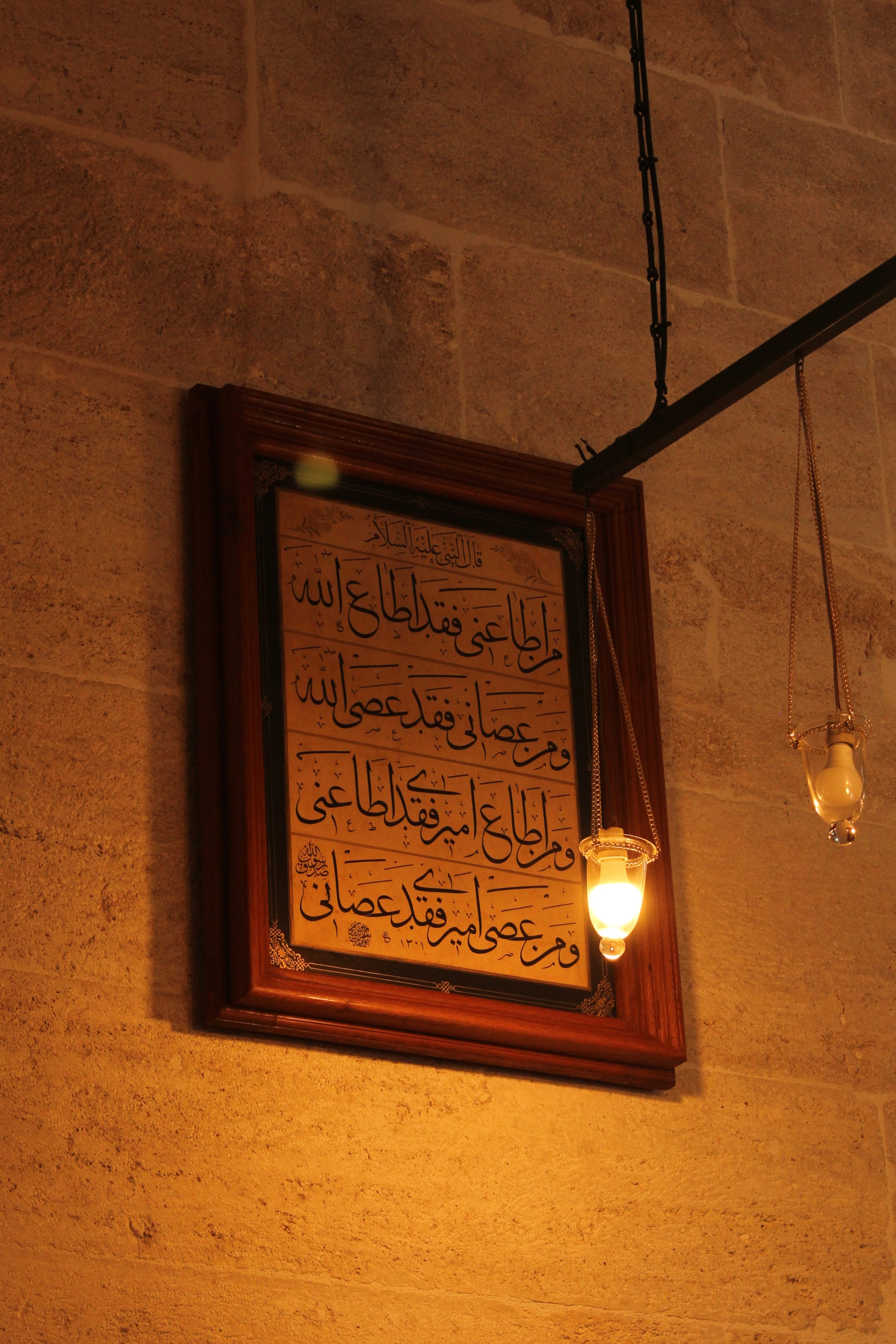 Framed Arabic calligraphy illuminated by soft lighting, set against a stone wall indoors.