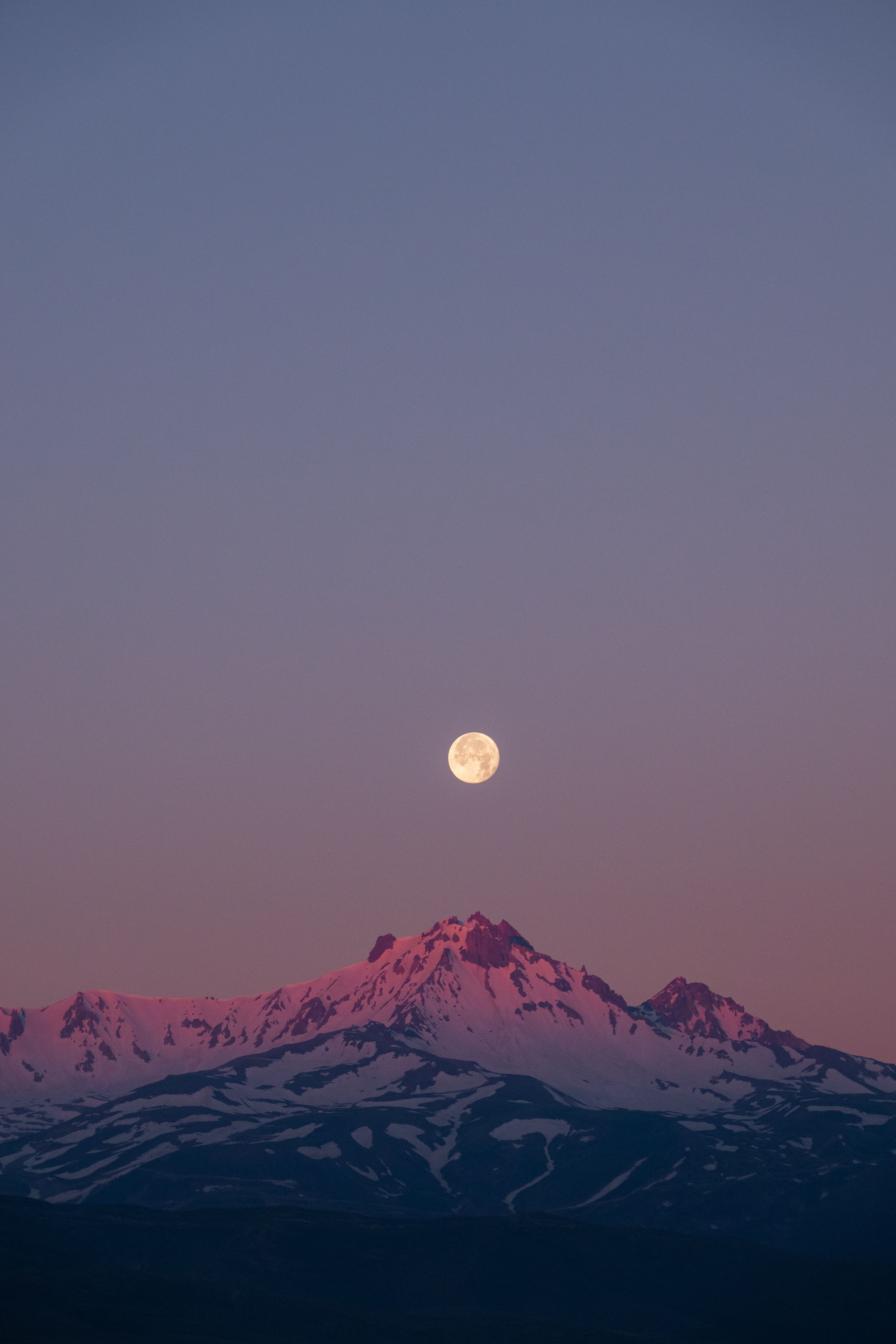 Full moon rising above snow-covered mountain peaks at twilight, showcasing a serene natural landscape.