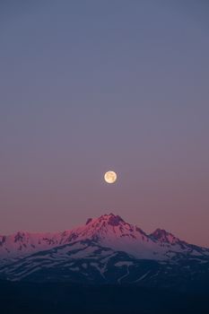 Full moon rising above snow-covered mountain peaks at twilight, showcasing a serene natural landscape.