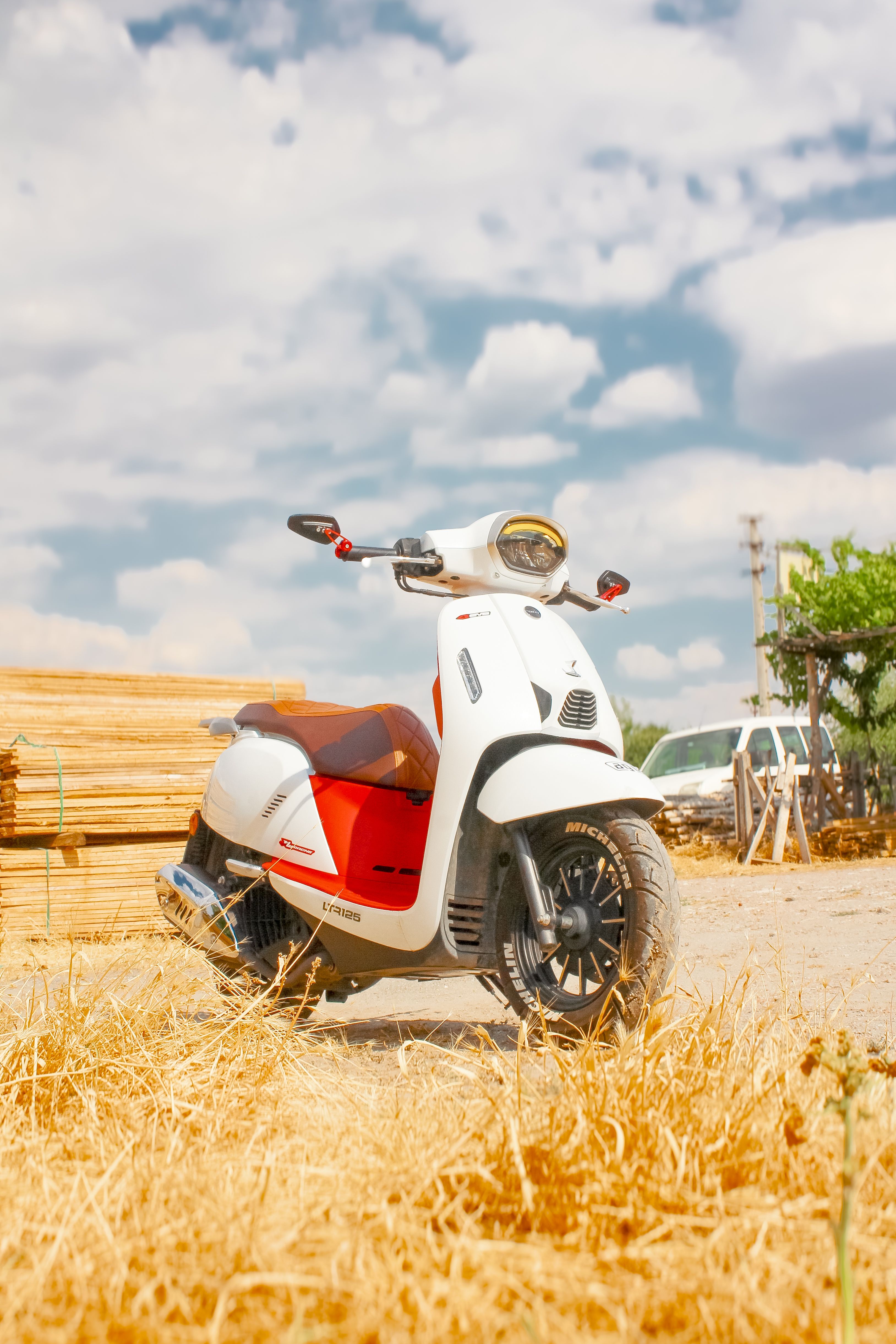 White scooter with red accents parked in a sunny outdoor setting next to lumber stacks.