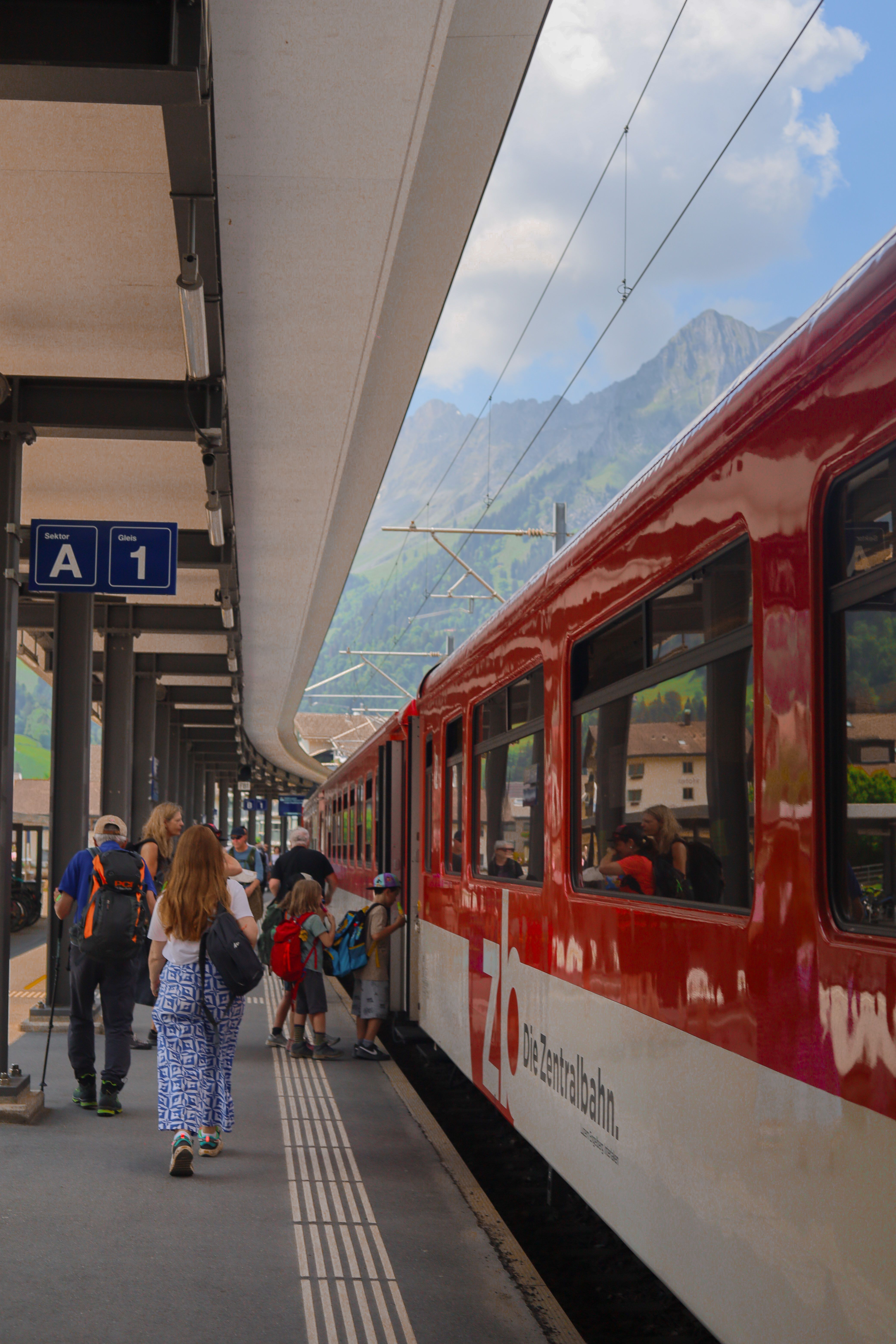 Passengers boarding a red Zentralbahn train at Engelberg station in Switzerland with scenic mountain backdrop.
