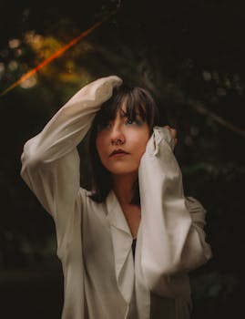 一位年轻女子身穿白色上衣，在户外若有所思地摆着姿势，双手放在头上