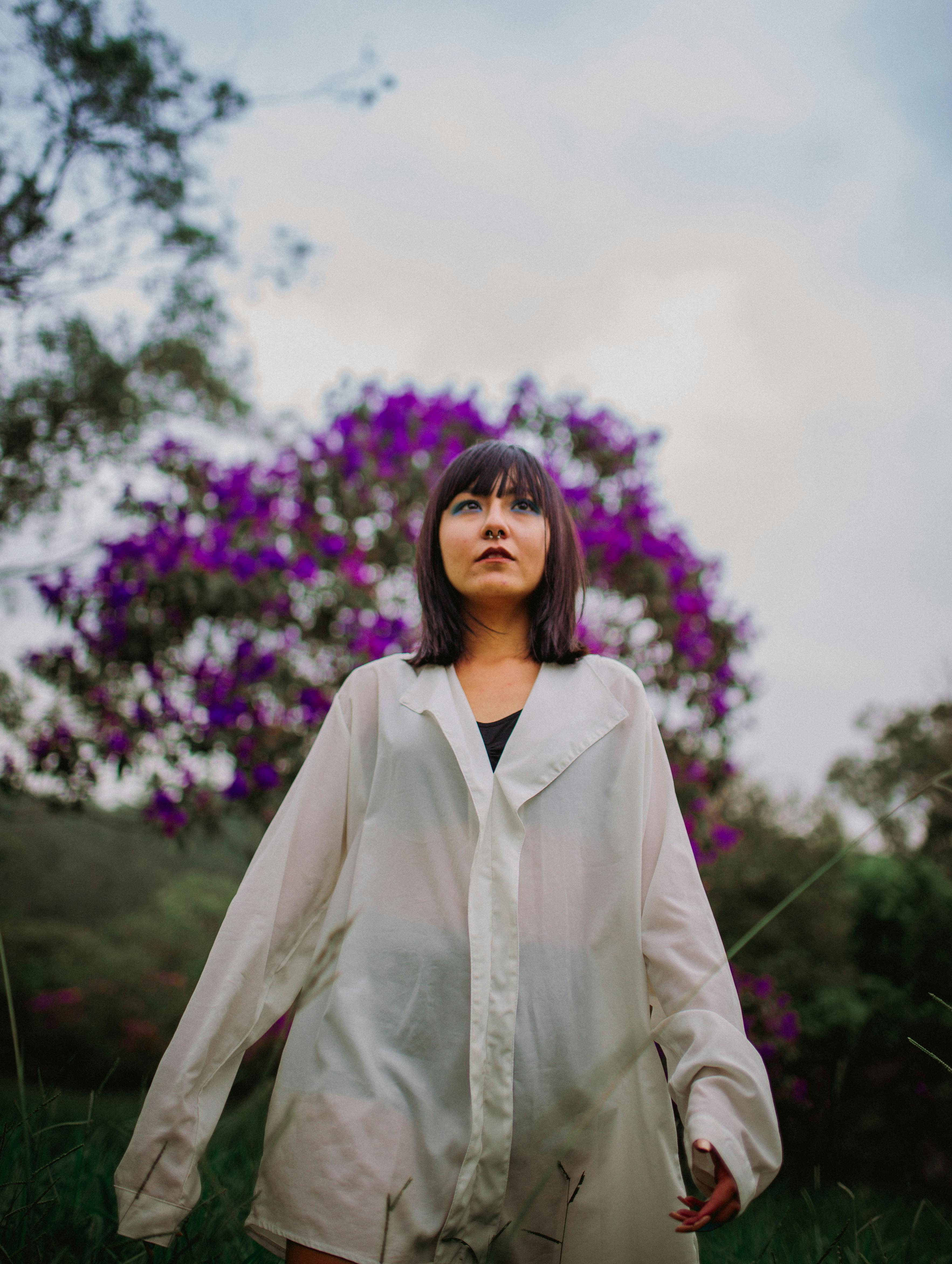 一位穿着白色衬衫的年轻女子站在户外，身后是紫色的花朵