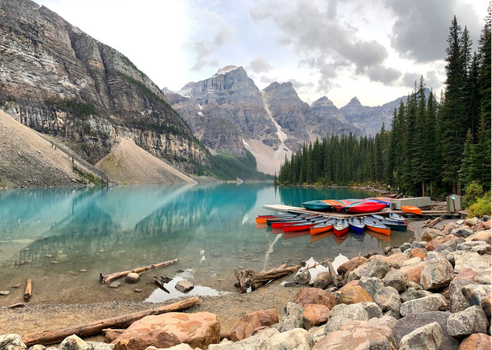 Beautiful scene of Moraine Lake with canoes and surrounding Rocky Mountains under a cloudy sky.