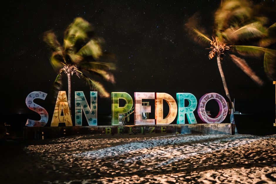 Colorful San Pedro Belize sign illuminated under a starry night sky with palm trees and sandy beach.