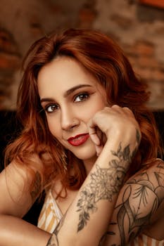 Portrait of positive young tattooed with ginger hair leaning on hand and looking at camera
