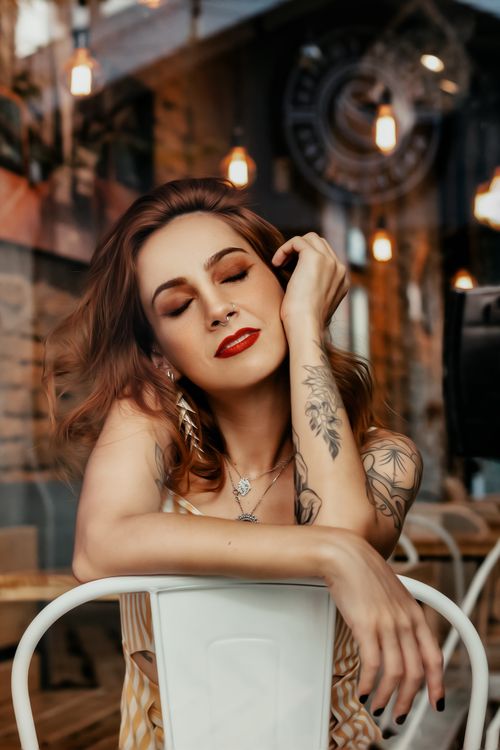 Free Delighted young redhead tattooed lady in stylish outfit sitting on chair with closed eyes and enjoying summer day Stock Photo