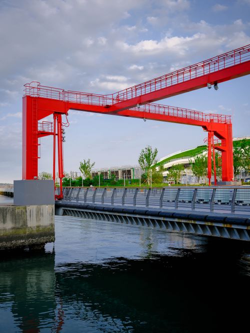 Free Striking red crane structure spanning a serene waterway, surrounded by greenery under a partly cloudy sky. Stock Photo