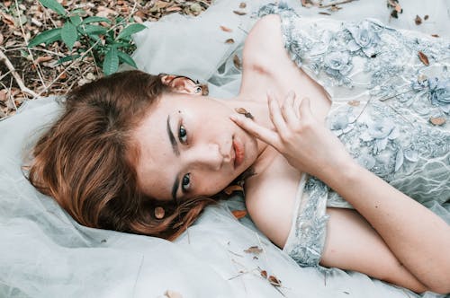 From above of young romantic ethnic female in elegant dress lying on white blanket on ground in forest and looking at camera