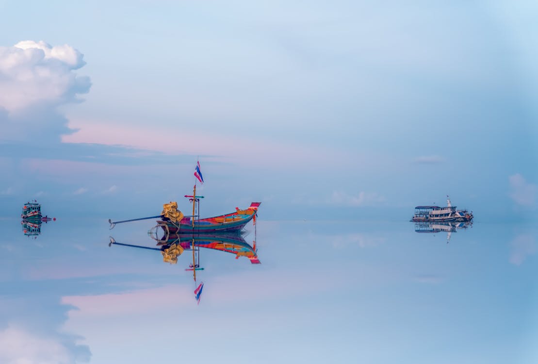 Traditional colorful fishing boat floating on surface of calm sea near modern vessels against sunset sky reflecting in blue water