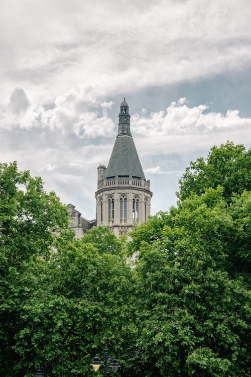 Free Gothic-style tower surrounded by lush trees under a cloudy sky, blending architecture and nature. Stock Photo